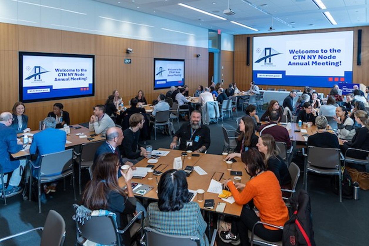 Meeting participants seated around tables looking at slides on two screens
